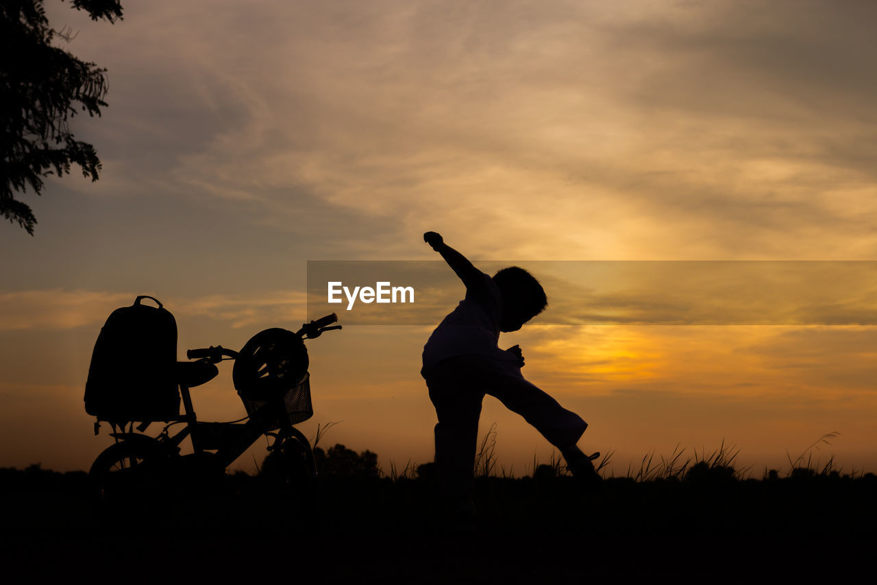 Silhouette boy playing by bicycle on land against sky during sunset