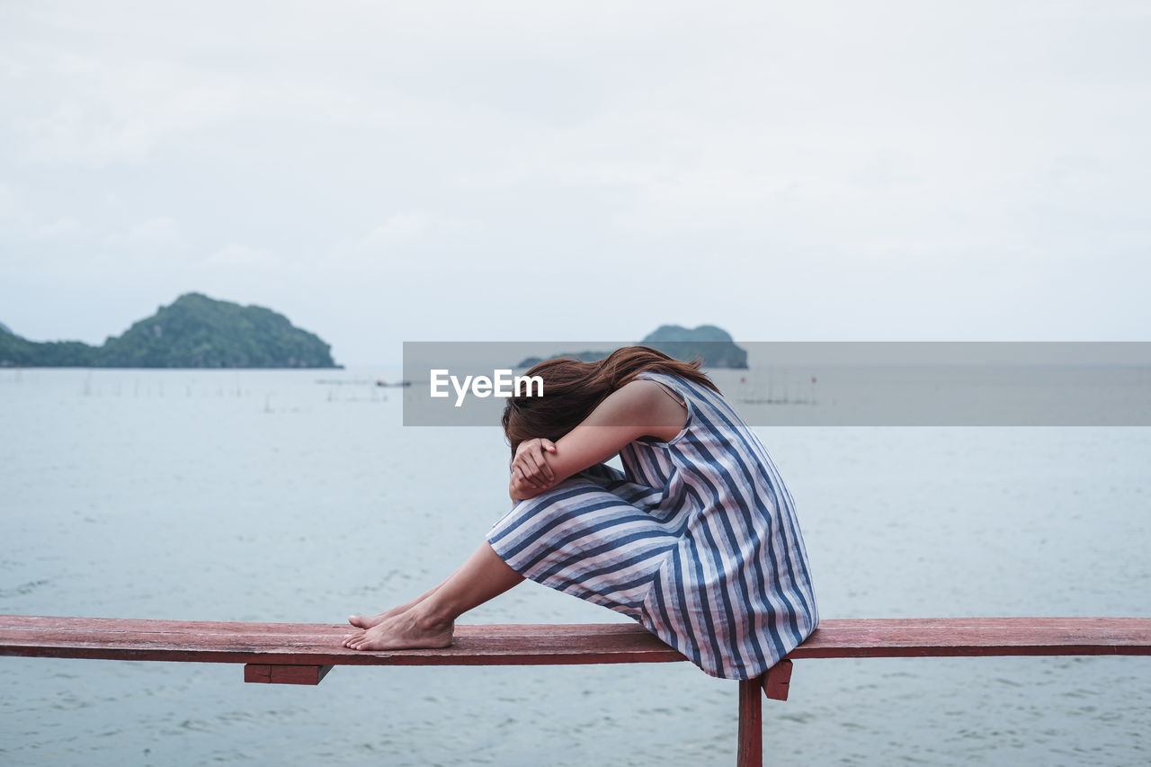 Woman sitting on railing by sea against sky