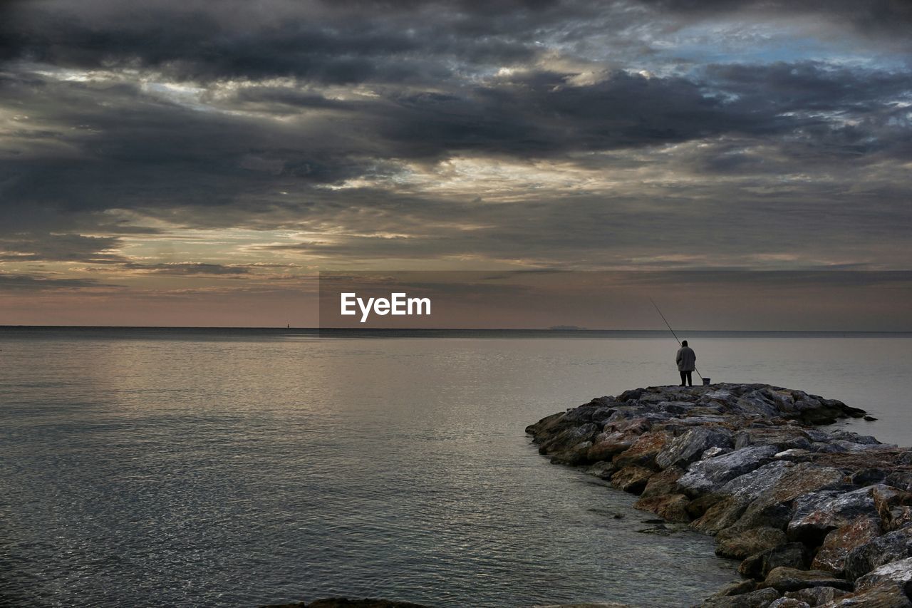 Distant view of man standing on rocks by sea against cloudy sky during sunset