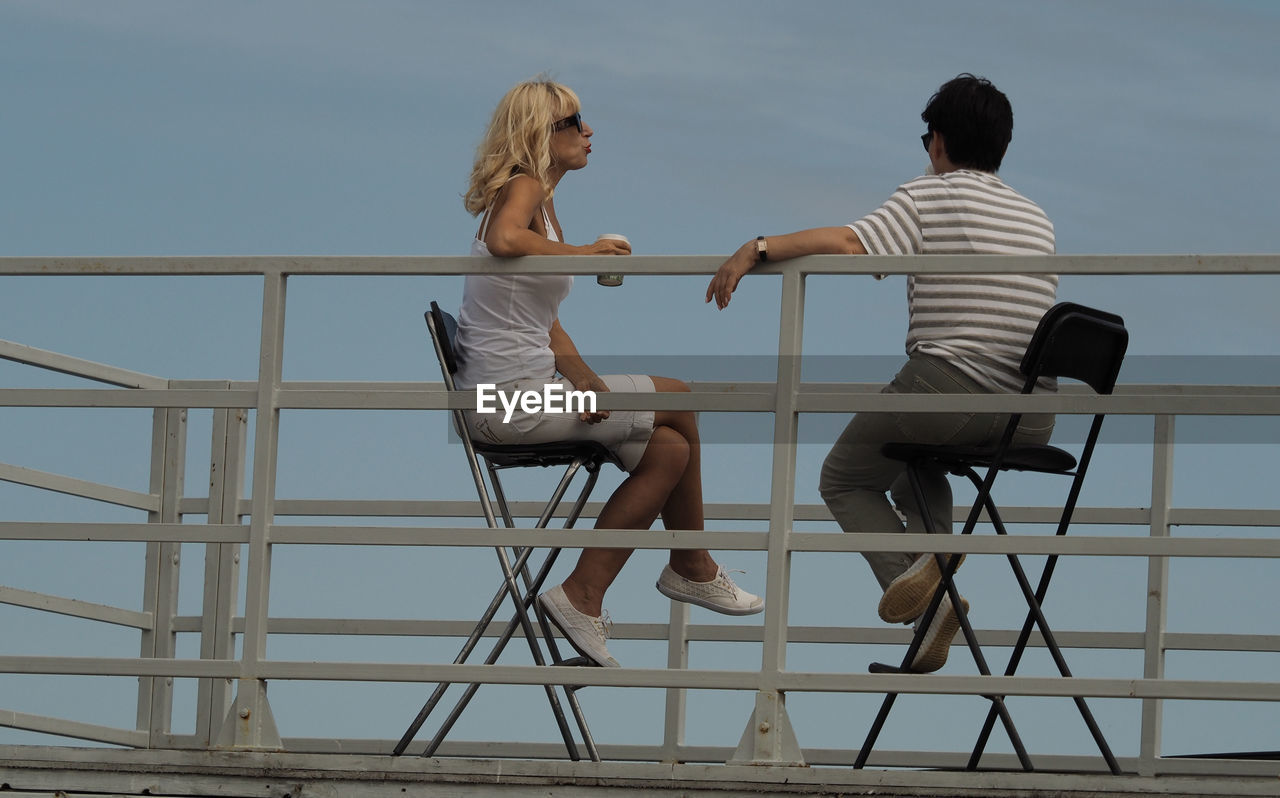 two people, adult, togetherness, women, men, sitting, full length, casual clothing, bonding, positive emotion, female, emotion, sky, leisure activity, love, young adult, nature, railing, lifestyles, architecture, friendship, day, seat, relaxation, water, outdoors, rear view, happiness, communication, enjoyment, romance, furniture, chair