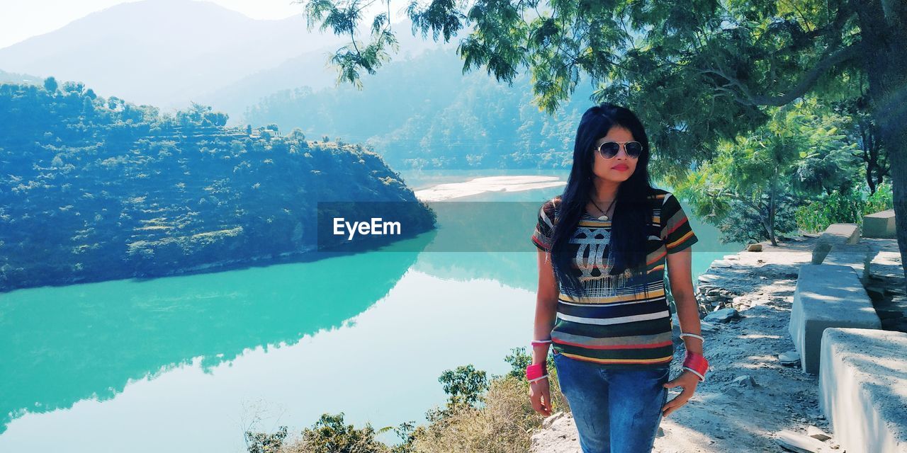 PORTRAIT OF BEAUTIFUL YOUNG WOMAN STANDING BY TREE AGAINST MOUNTAIN