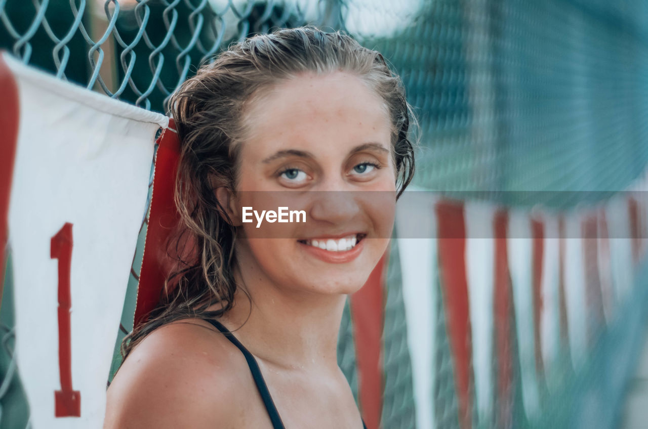 Portrait of smiling young woman by chainlink fence outdoors