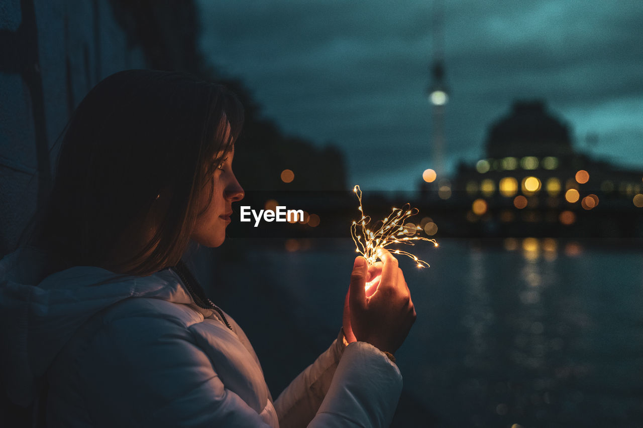 Young woman holding illuminated string lights against river at night