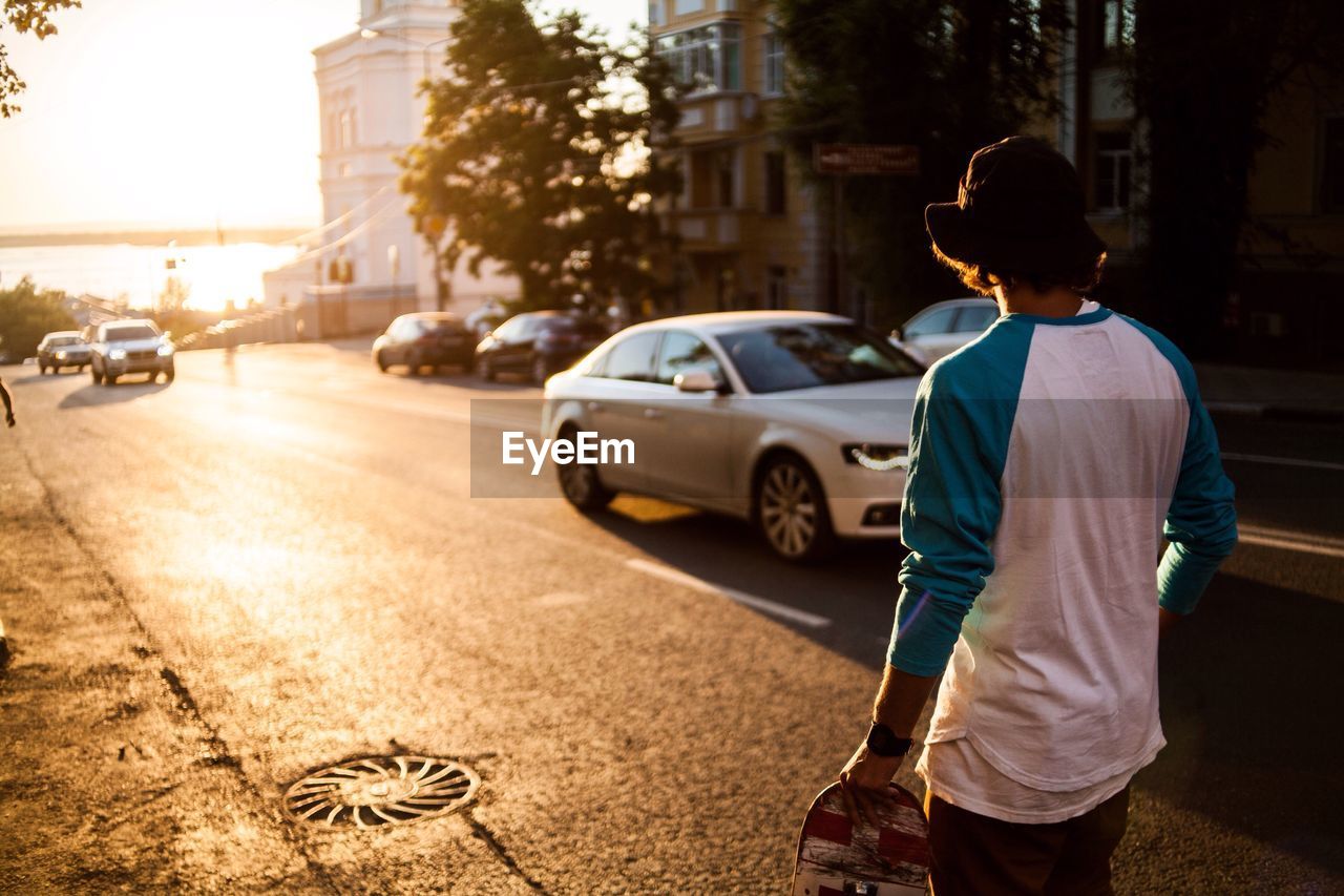 Rear view of man carrying skateboard while walking on road during sunset