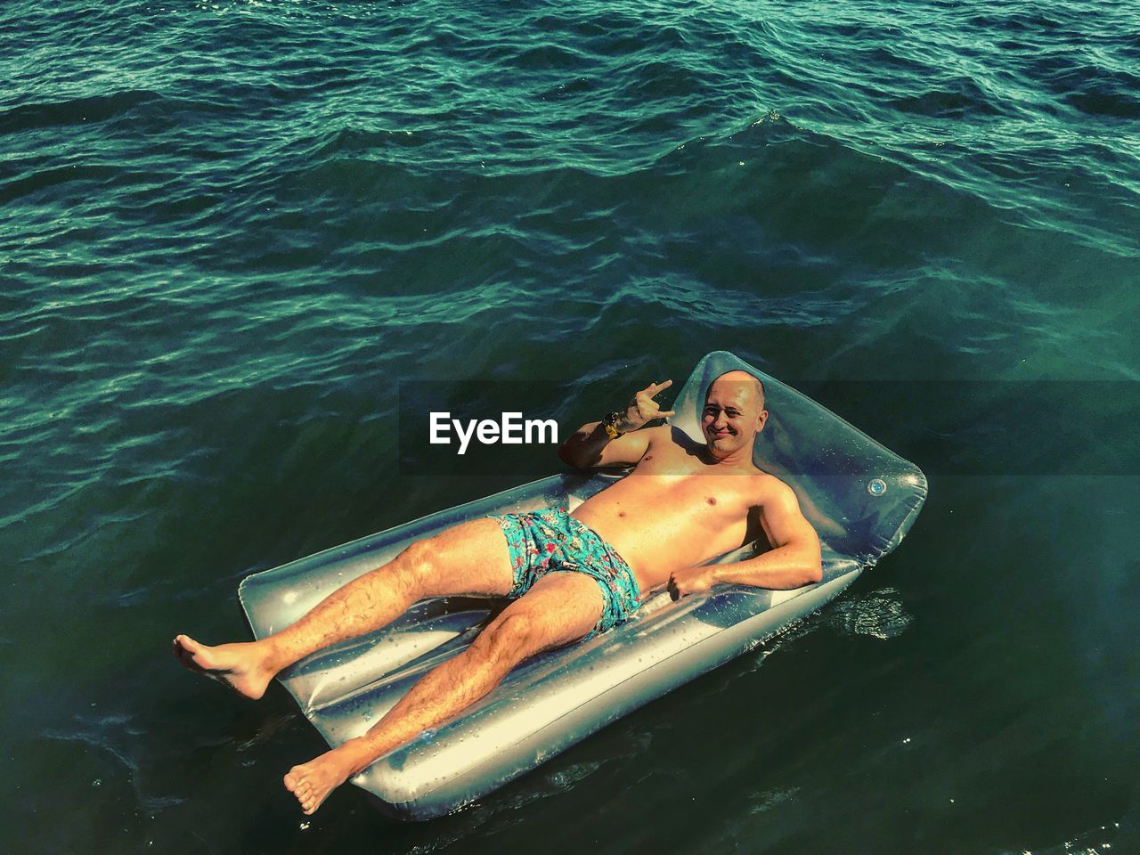 High angle portrait of shirtless mid adult man gesturing while lying on pool raft in sea