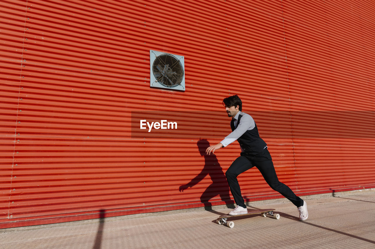 Businessman with longboard skateboarding on footpath by wall