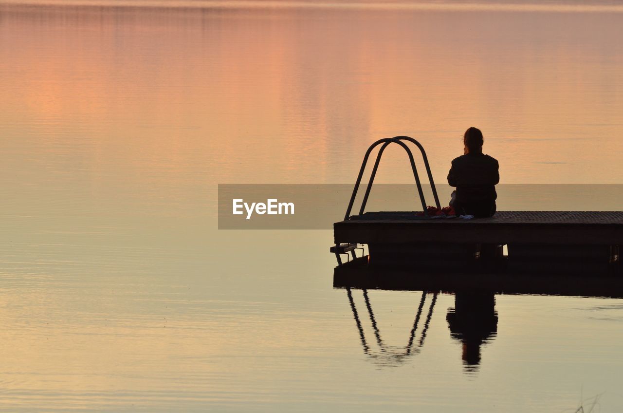 Rear view of woman sitting on pier over lake during sunset