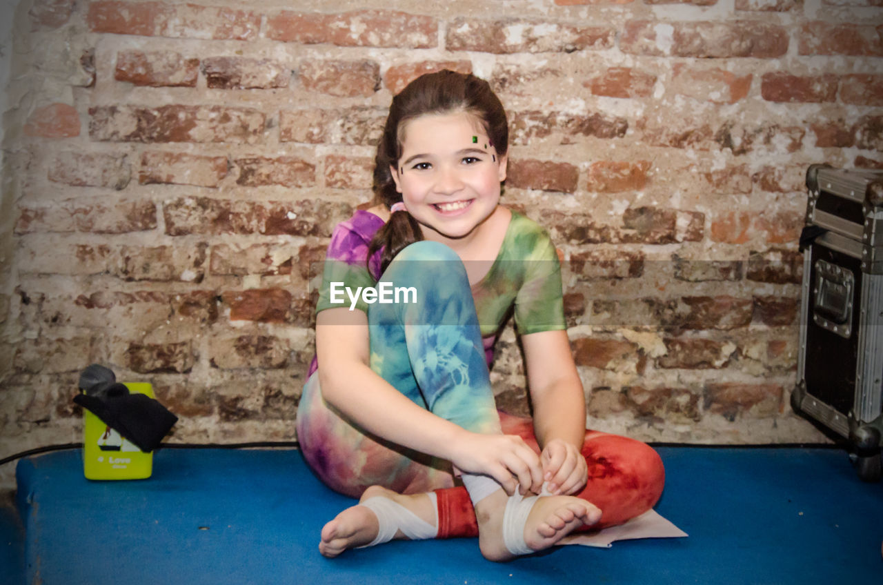 Portrait smiling girl sitting on floor against brick wall
