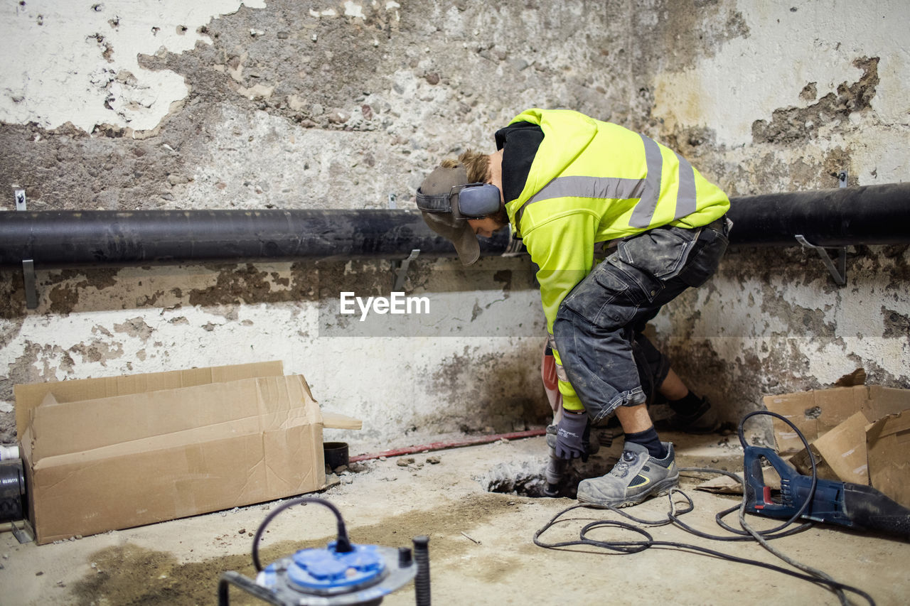 Concentrated plumber drilling floor by pipes in basement
