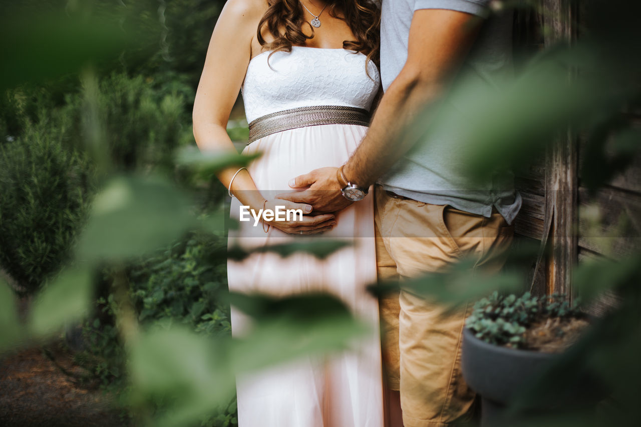 Midsection of man holding pregnant wife while standing by plants