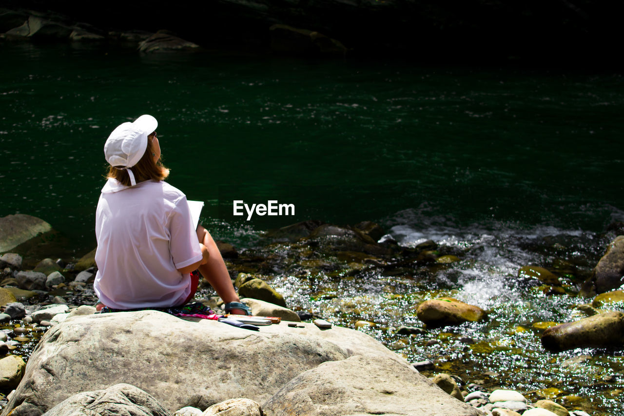 Rear view of girl sitting on rock by river