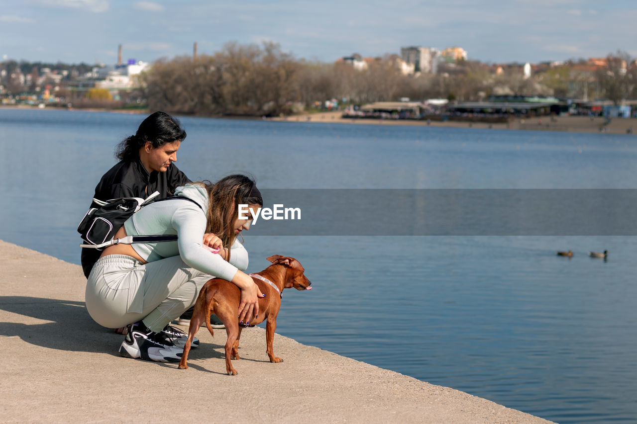 Young girl and her mother on relaxing vacation by the lake with their cute miniature pinscher.