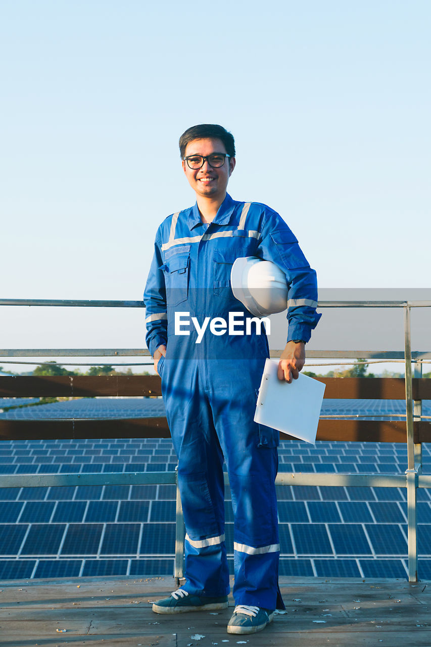Portrait of smiling electrician standing by railing against solar panels