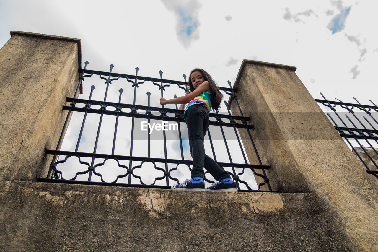 Low angle view of carefree girl standing on retaining wall by railing against sky