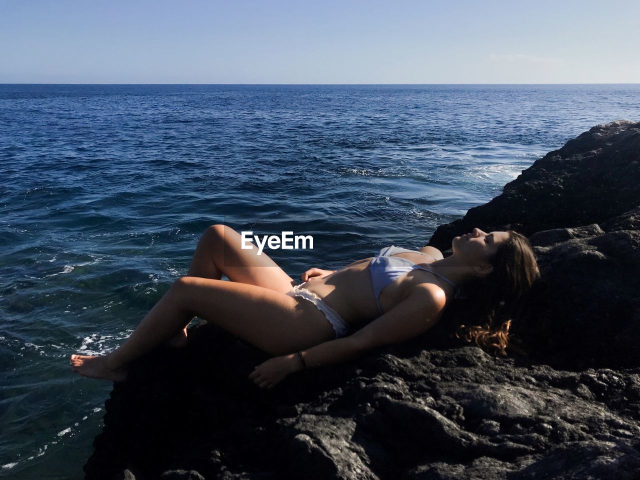 Woman in bikini relaxing on rock by sea against sky