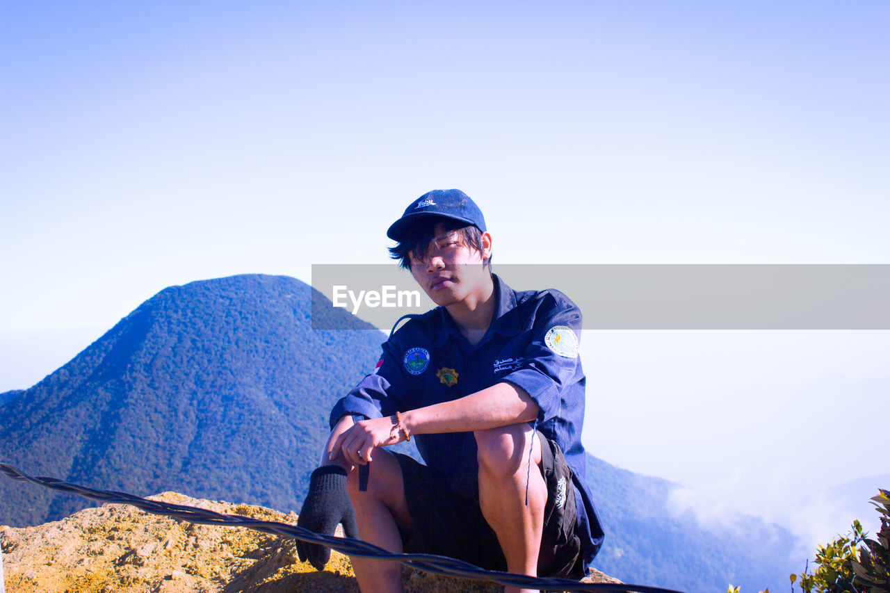 YOUNG MAN SITTING AGAINST MOUNTAINS AGAINST CLEAR BLUE SKY