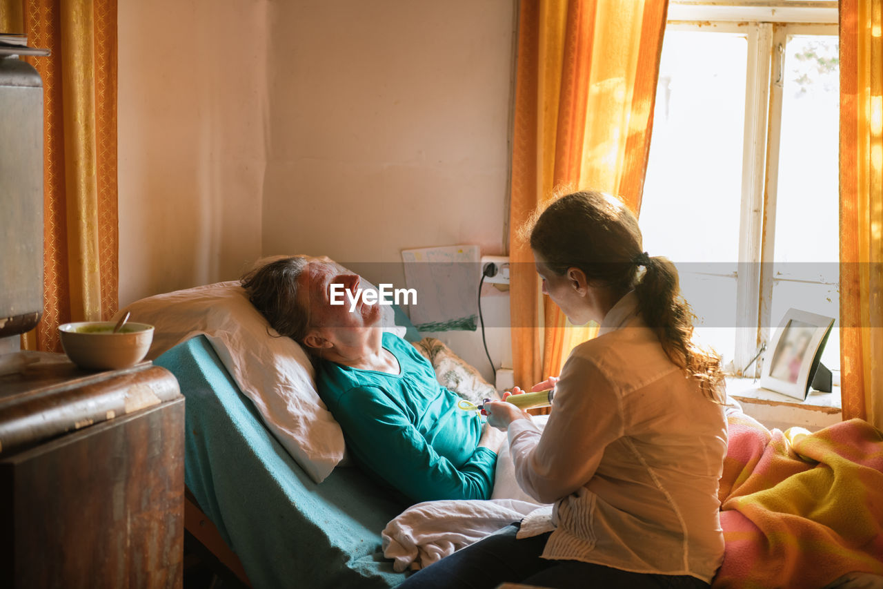 Female nurse feeding paralyzed elderly lady with puree through tube and syringe while taking care of patient at home