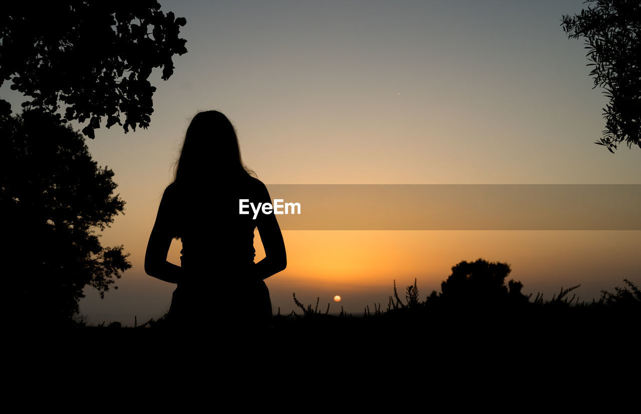 SILHOUETTE WOMAN STANDING BY TREE ON FIELD AGAINST SKY AT SUNSET