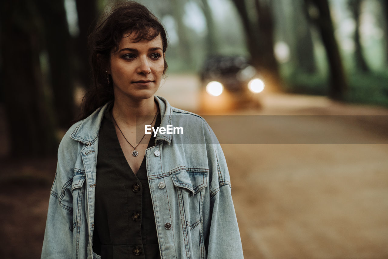 Young woman standing alone on a forest road with a car behind