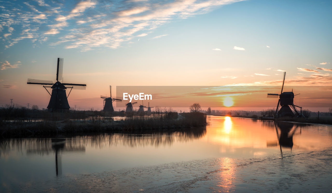 Silhouette windmills by lake against sky during sunset