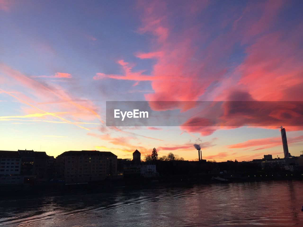 Silhouette buildings by river against sky at sunset