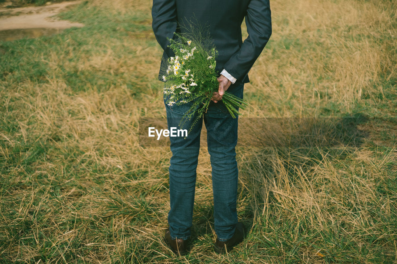 Low section of bridegroom holding bouquet while standing on grassy field in park