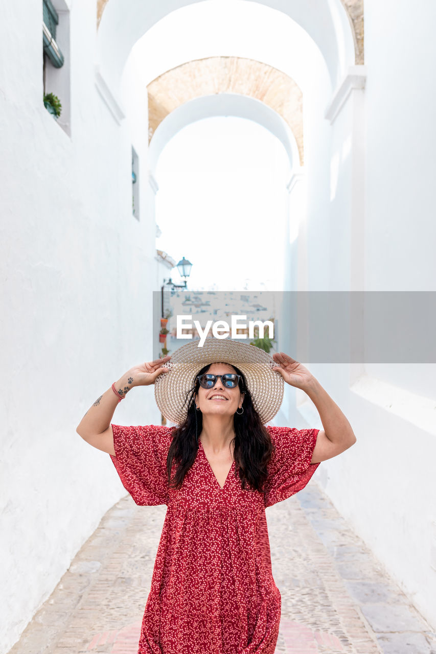 Front view of female tourist with raised arms and hat on narrow street between old houses in greece