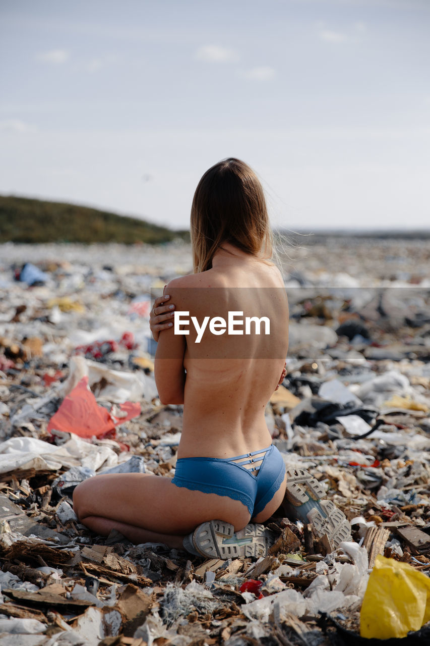 Rear view of shirtless woman sitting on garbage against sky