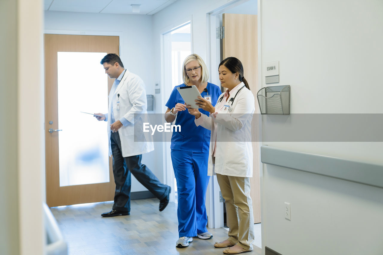 Female doctors discussing over tablet computer while male colleague walking by in hospital