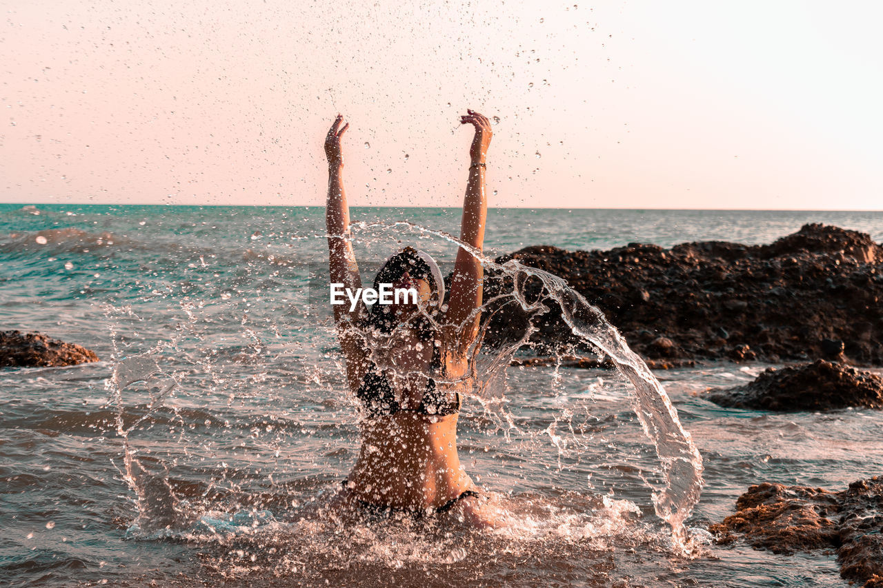 Woman splashing water in sea against clear sky at sunset