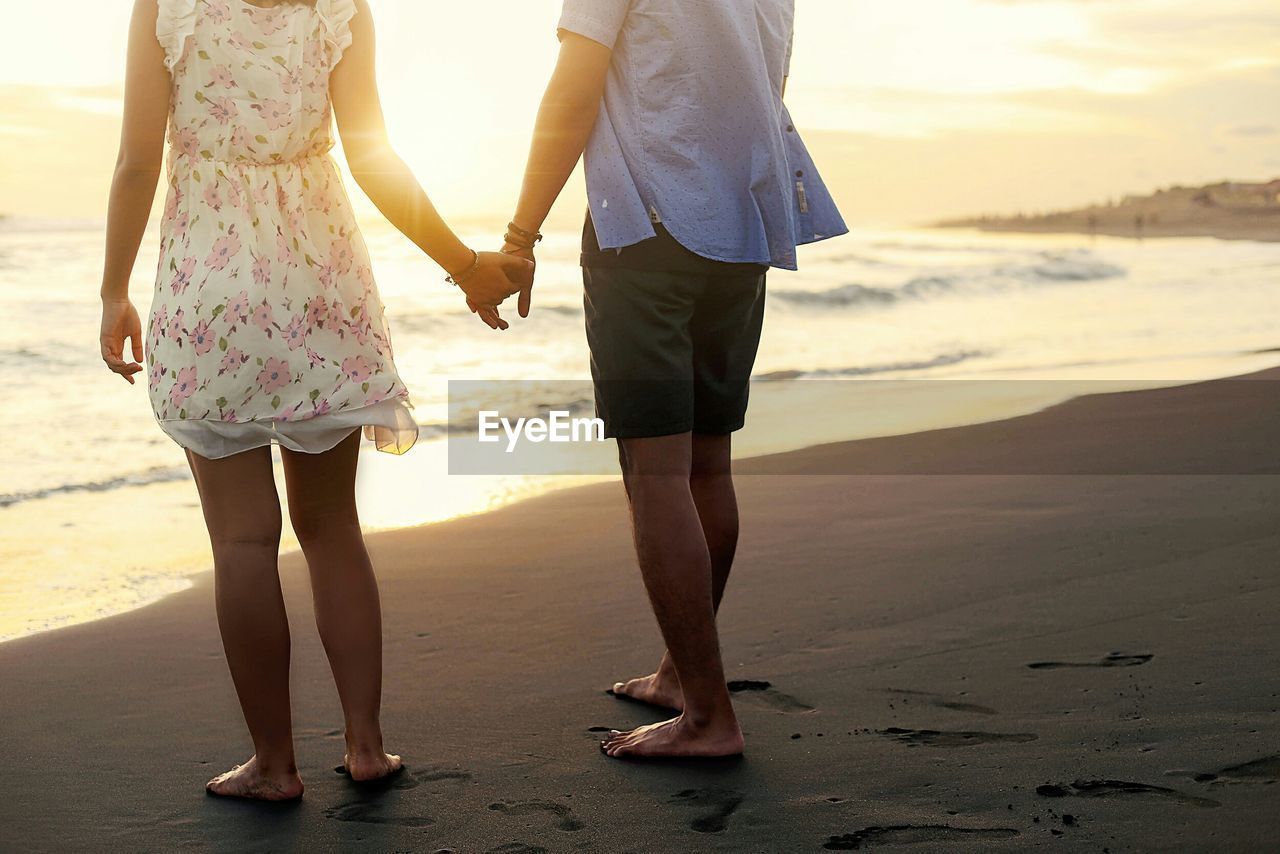 Low section of couple holding hands while standing on beach