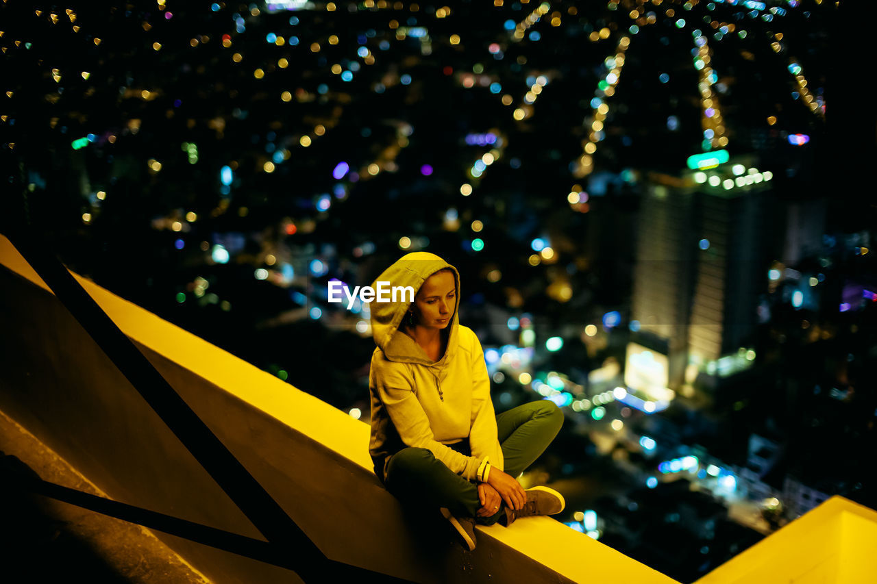 Woman sitting on retaining wall against illuminated city at night