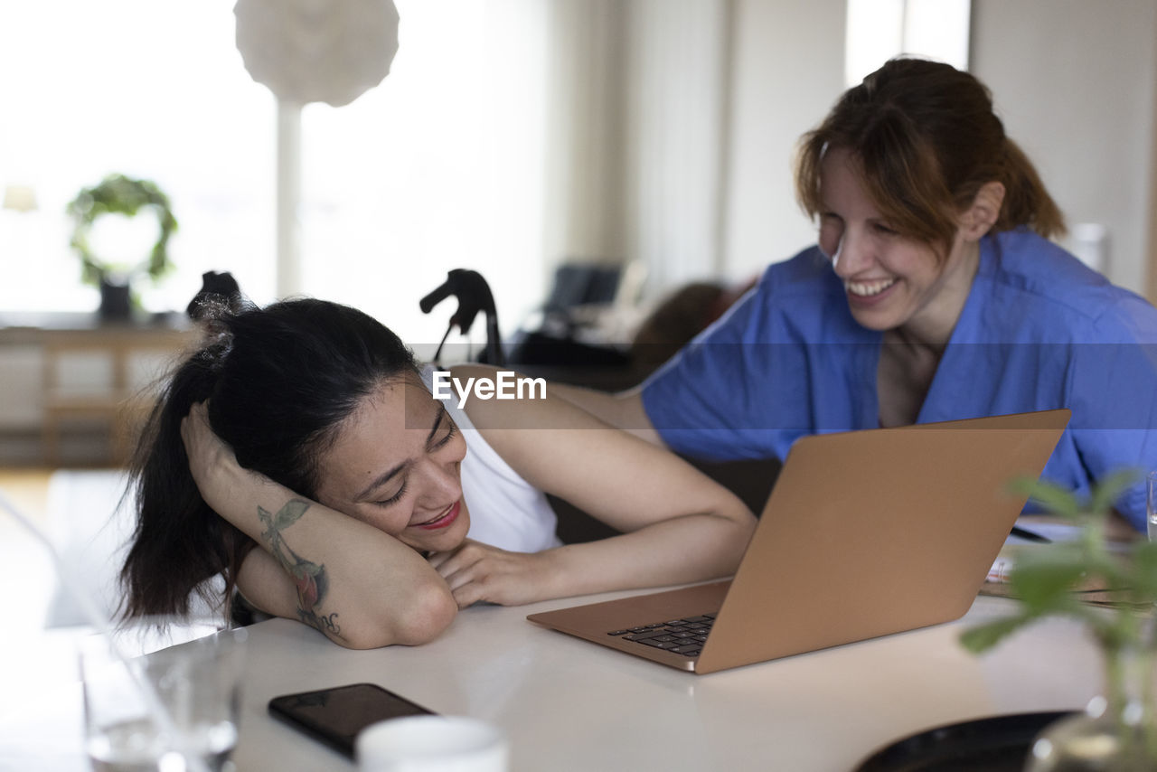 Happy female caregiver talking to woman with paraplegia watching laptop at home