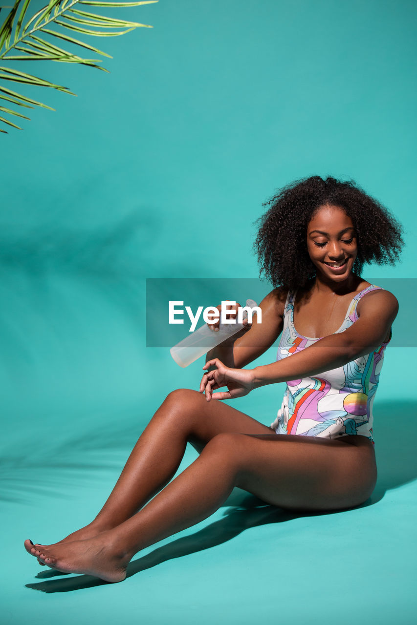 Full body feminine young african american female in one piece swimwear applying suntan spray on smooth skin while sitting on blue studio background