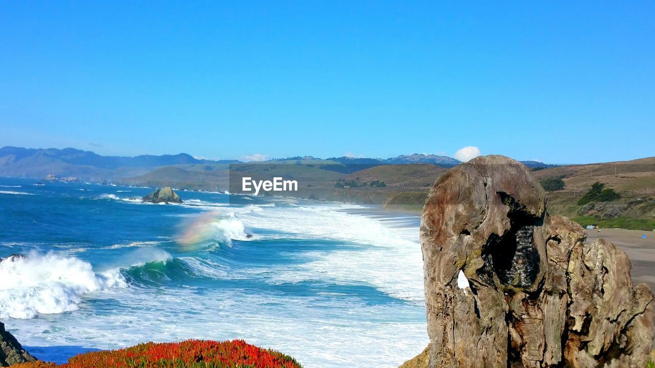 Scenic view of rainbow waves in the sea against clear blue sky and cliffs