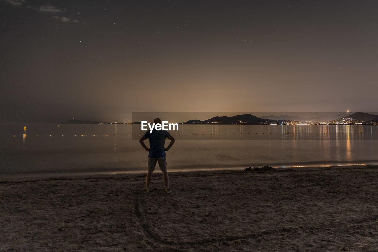 Rear view of man standing at beach against sky at night