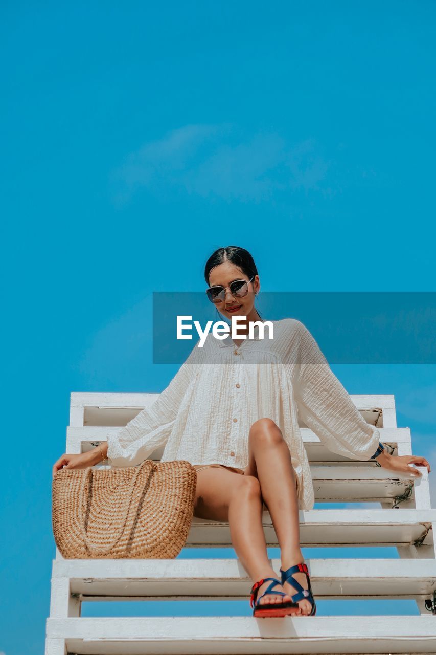 Portrait of young woman sitting on sunglasses against blue sky