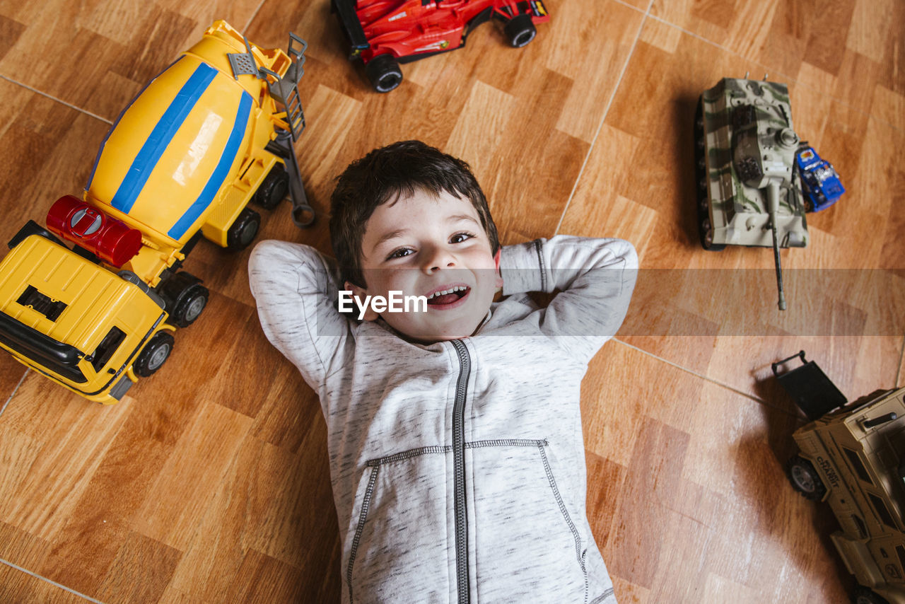 From above of cheerful little boy lying on parquet floor and looking at camera while playing with toy cars at home
