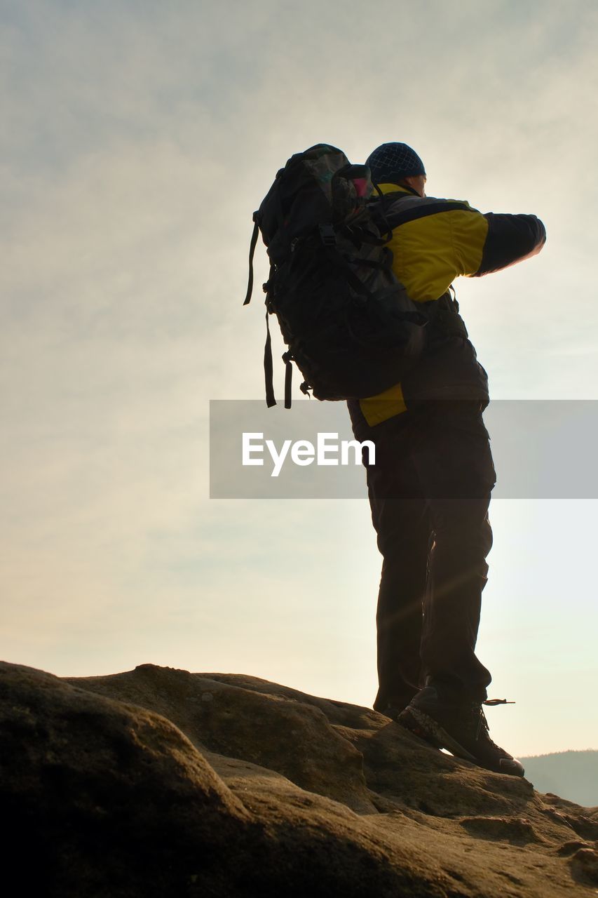 Tourist with backpack and poles in hands stand on cliff and watching into deep misty valley bellow.