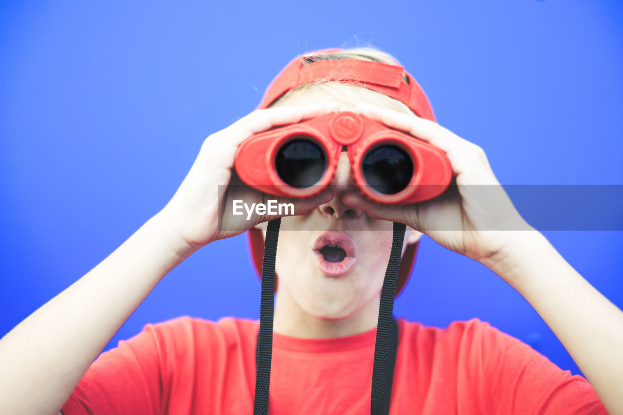 Close-up of boy looking through binoculars while standing against blue background