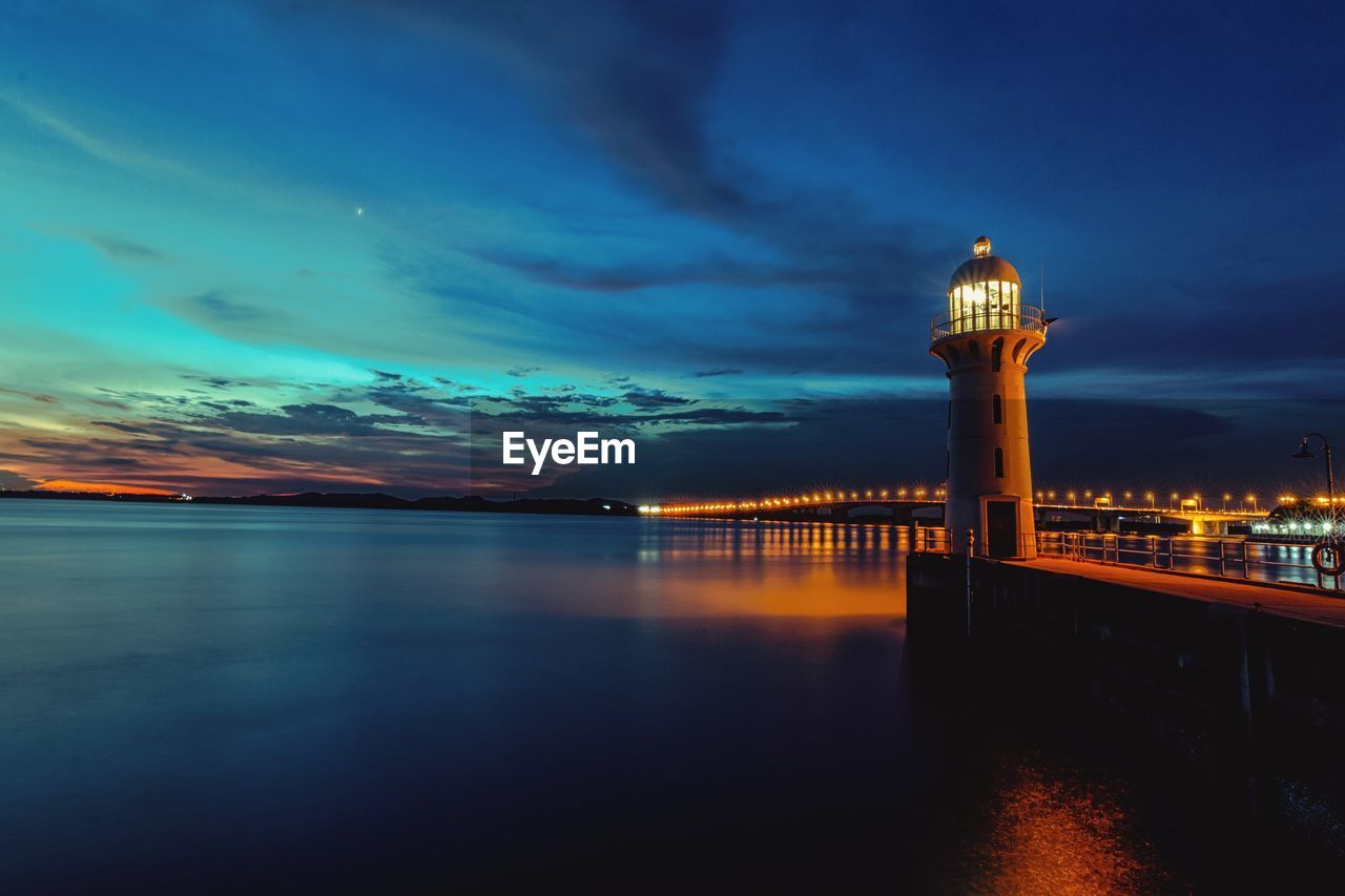 Illuminated lighthouse by sea against cloudy sky at dusk