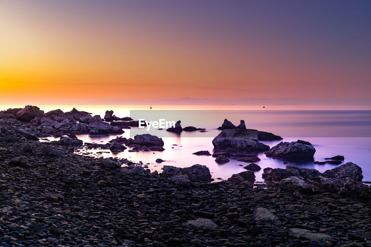 SCENIC VIEW OF ROCKS AT BEACH AGAINST SKY DURING SUNSET