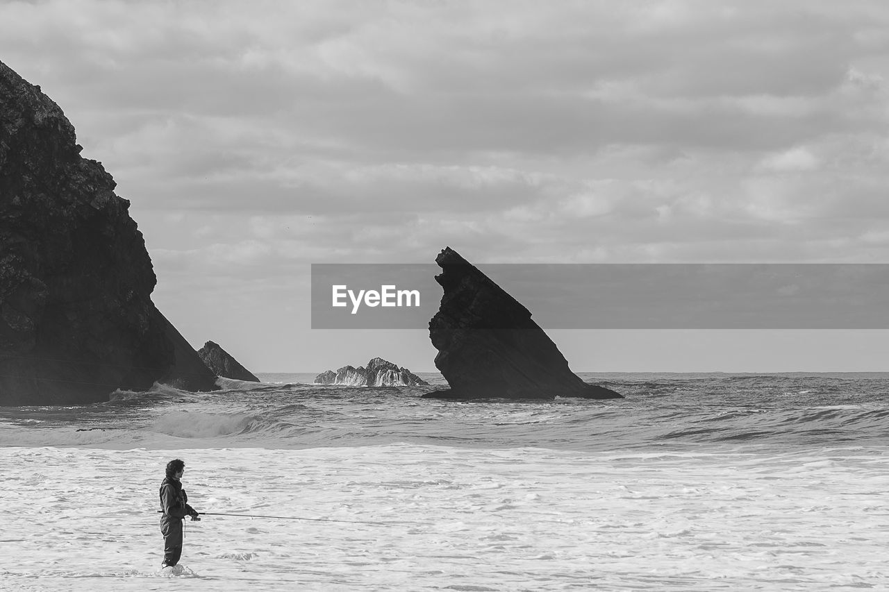 Man on the ocean fishing against sky and rocks 