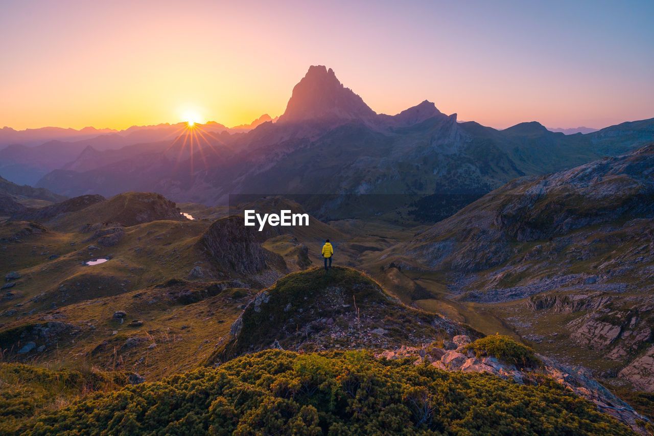 Distant unrecognizable tourists standing on rocky formation against rough mountain range during trip in nature of spain against cloudless sky