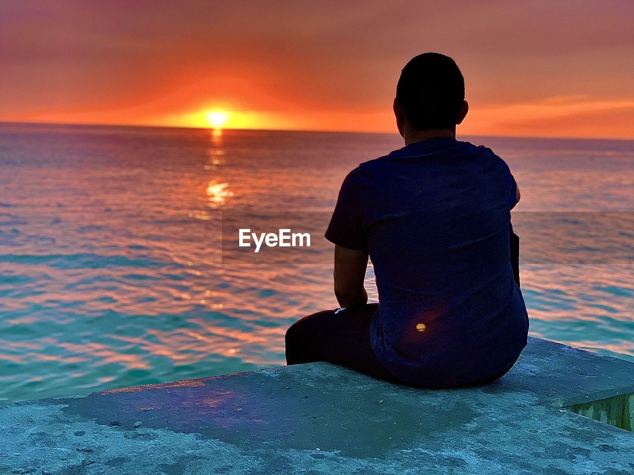 Rear view of man sitting on sea against sky during sunset