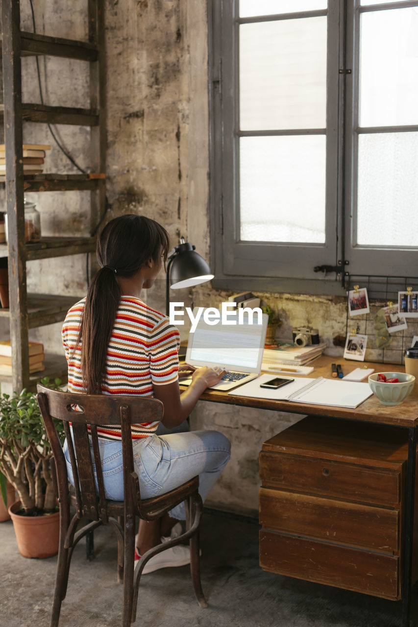 Back view of young woman sitting at desk in a loft working on laptop