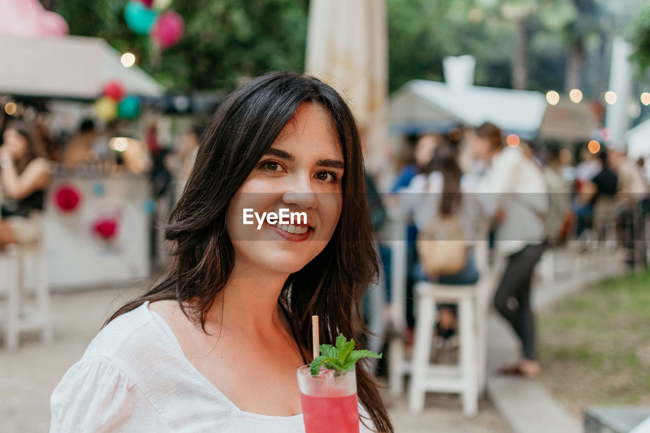 Beautiful young woman drinking strawberry mojito cocktail in park in city