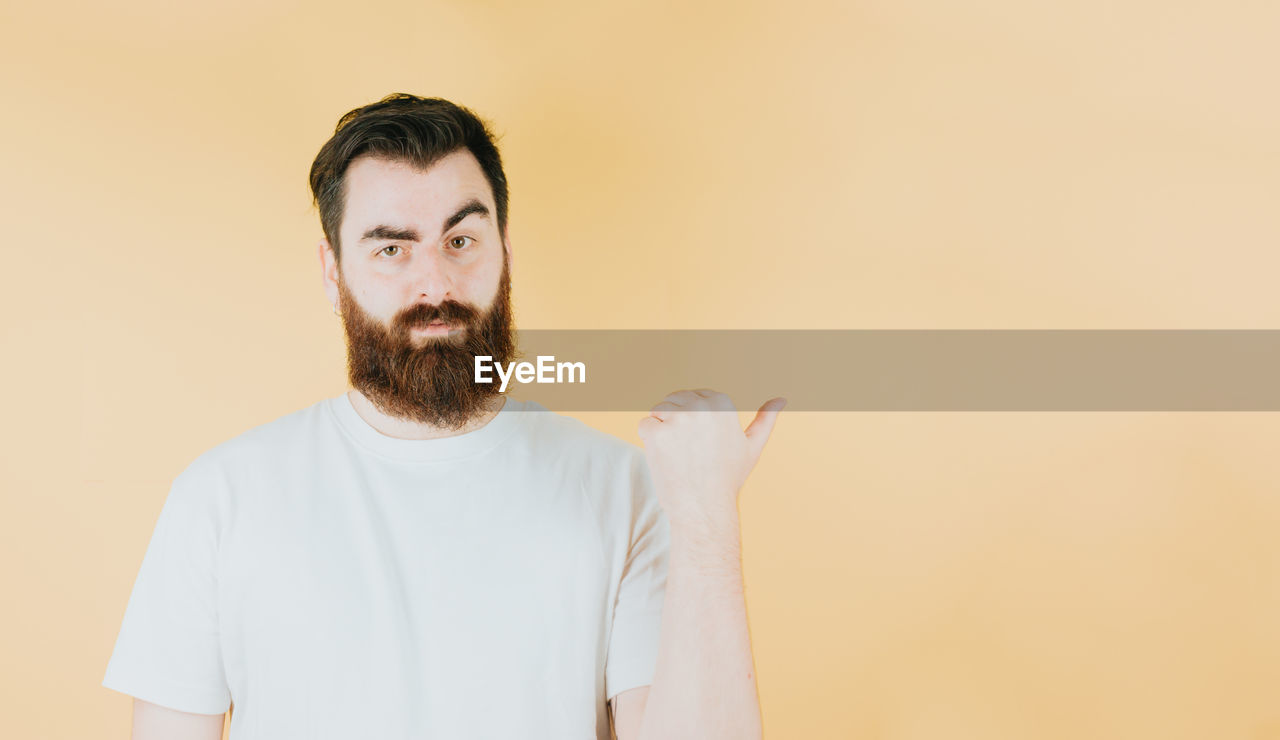 beard, facial hair, one person, adult, portrait, indoors, men, studio shot, copy space, person, colored background, waist up, front view, looking at camera, clothing, standing, human hair, brown hair, young adult, moustache, human face, casual clothing, looking, headshot, lifestyles, emotion, contemplation, beige background