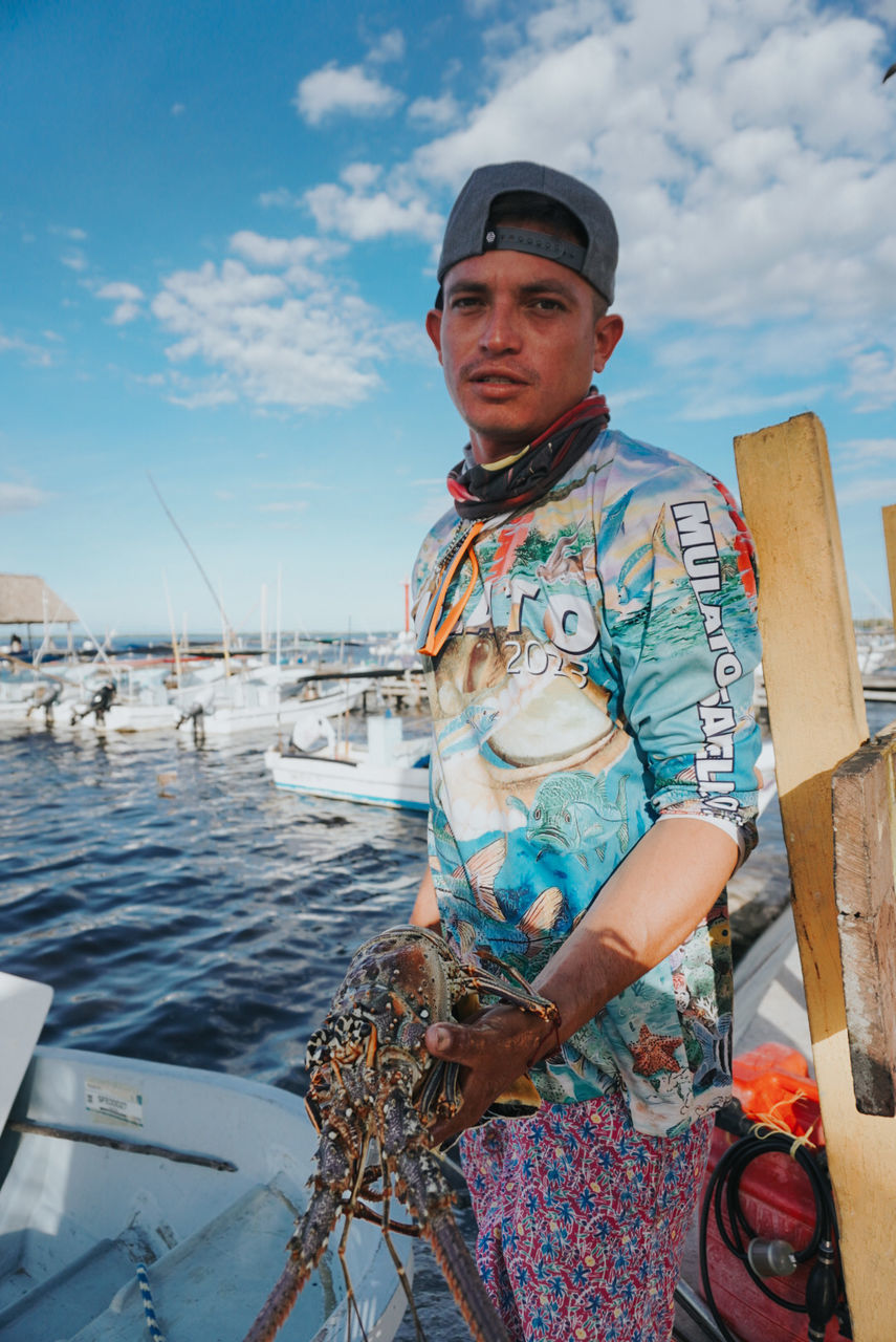 one person, water, sky, adult, nautical vessel, sea, transportation, nature, standing, looking at camera, cloud, portrait, occupation, day, men, blue, three quarter length, mode of transportation, vacation, fishing, ship, clothing, person, vehicle, sailor, travel, outdoors, front view, waist up, fishing industry, hat, fashion, boat, holding, working