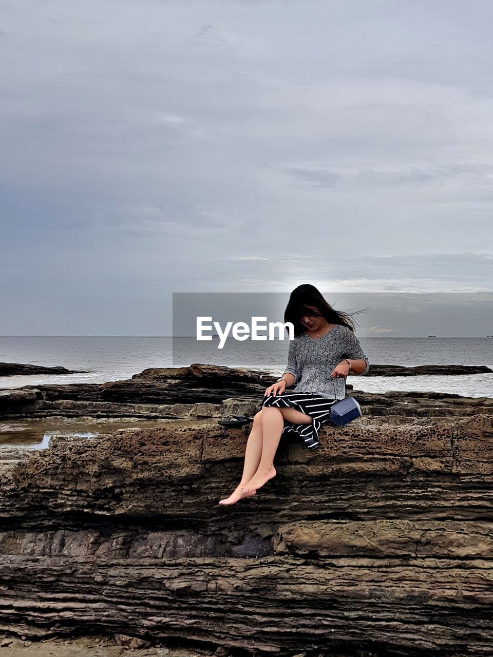 Woman sitting on rock formation by sea against cloudy sky